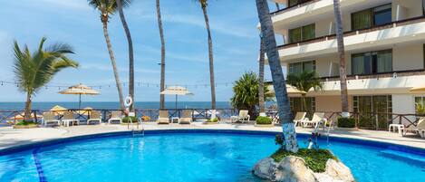 A pool with clear blue water, surrounded by palm trees and lounge chairs, with a view of the ocean and a multi-story building in the background.