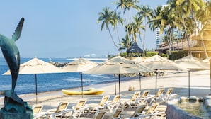Uma praia com cadeiras de praia, guarda-sóis e palmeiras, além de uma escultura.