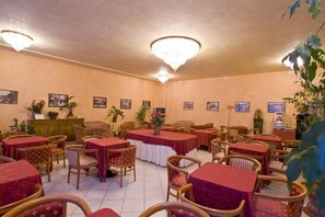 Un espace repas avec plusieurs tables et chaises, dont certaines sont recouvertes de nappes rouges. Des plantes en pot et des tableaux encadrés décorent les murs.