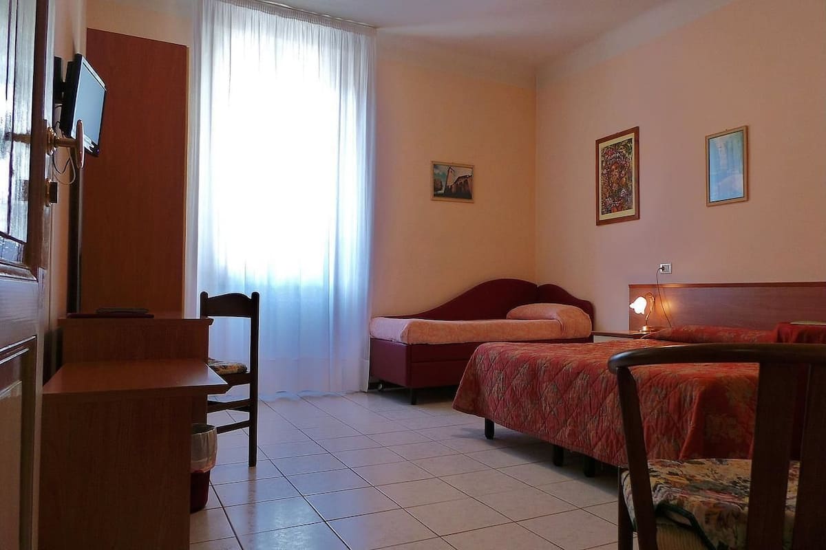 Ein Hotelzimmer mit einem Bett, einem Schreibtisch, einem Stuhl, einem an der Wand montierten Fernseher und einem Fenster mit Vorhängen.