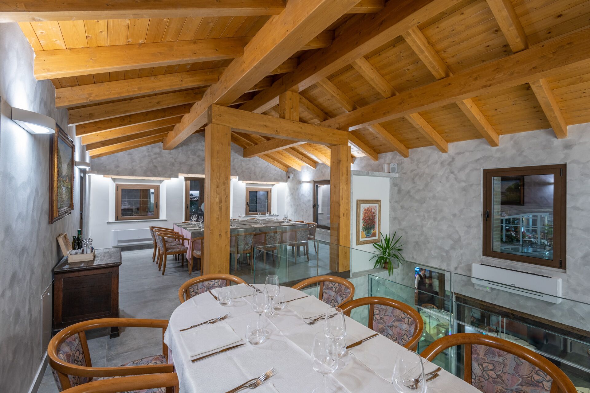Ein Essbereich mit einem langen Tisch, gedeckt für sechs Personen, Holzbalken an der Decke und Blick durch eine Glasabtrennung in einen weiteren Raum.
