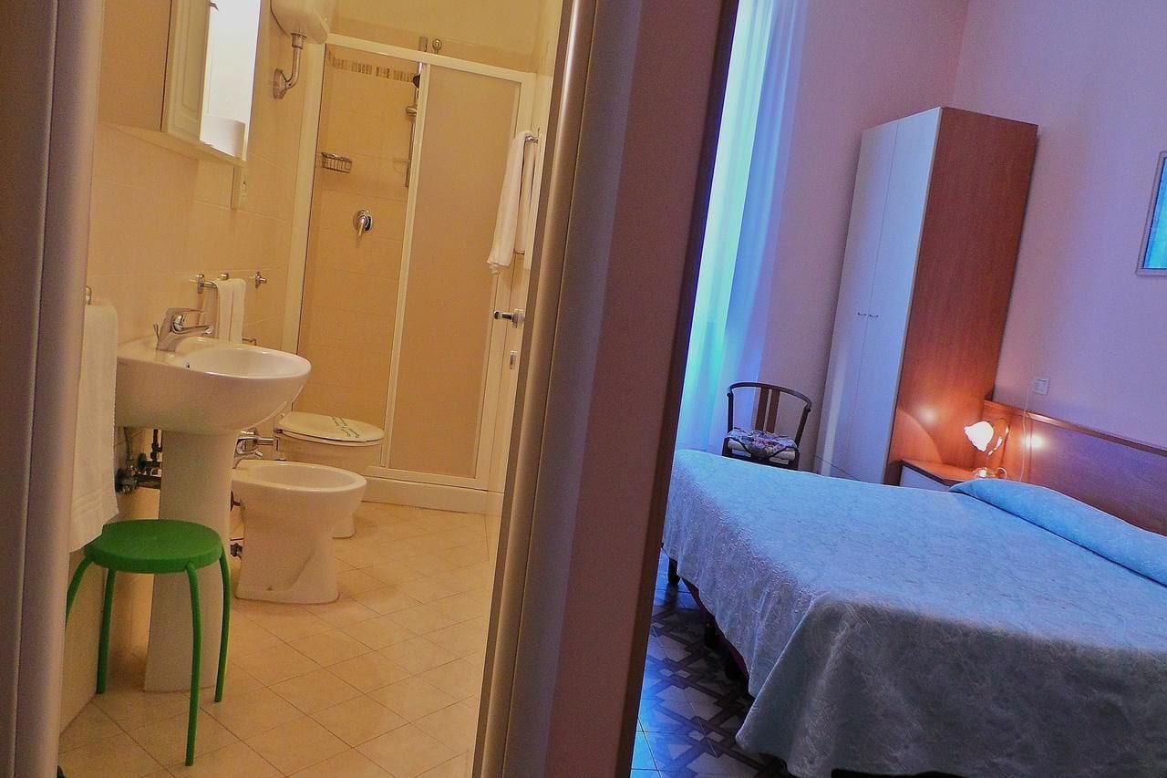 A bathroom with a sink, toilet, and shower. There is a green stool and a bed with a white bedspread in the adjacent room.