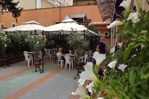Un espace café en plein air, avec des tables et des chaises sous des parasols, entouré de plantes et de fleurs en pot.