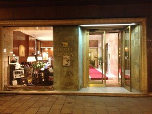 L’entrée d’un hôtel est marquée par un tapis rouge qui mène à l’intérieur, un comptoir d’accueil et une enseigne indiquant « HÔTEL ROMA ».