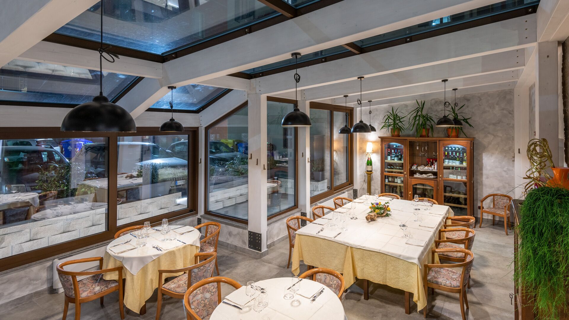 A modern dining area with high ceilings, skylights, and large windows offering views of the street. Tables are set with white tablecloths and wooden chairs.