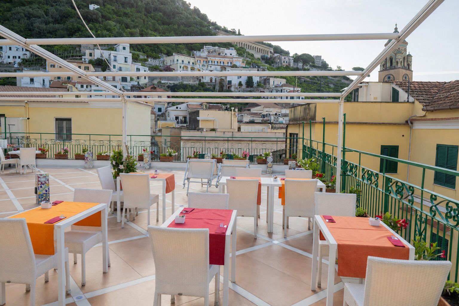 En udendørs terrasse med borde og stole, udsigt til en by på en bakke og et grønt metalgelænder.