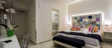 A modern hotel room with a bed, bedside table, chair, and a bathroom visible through an open door.