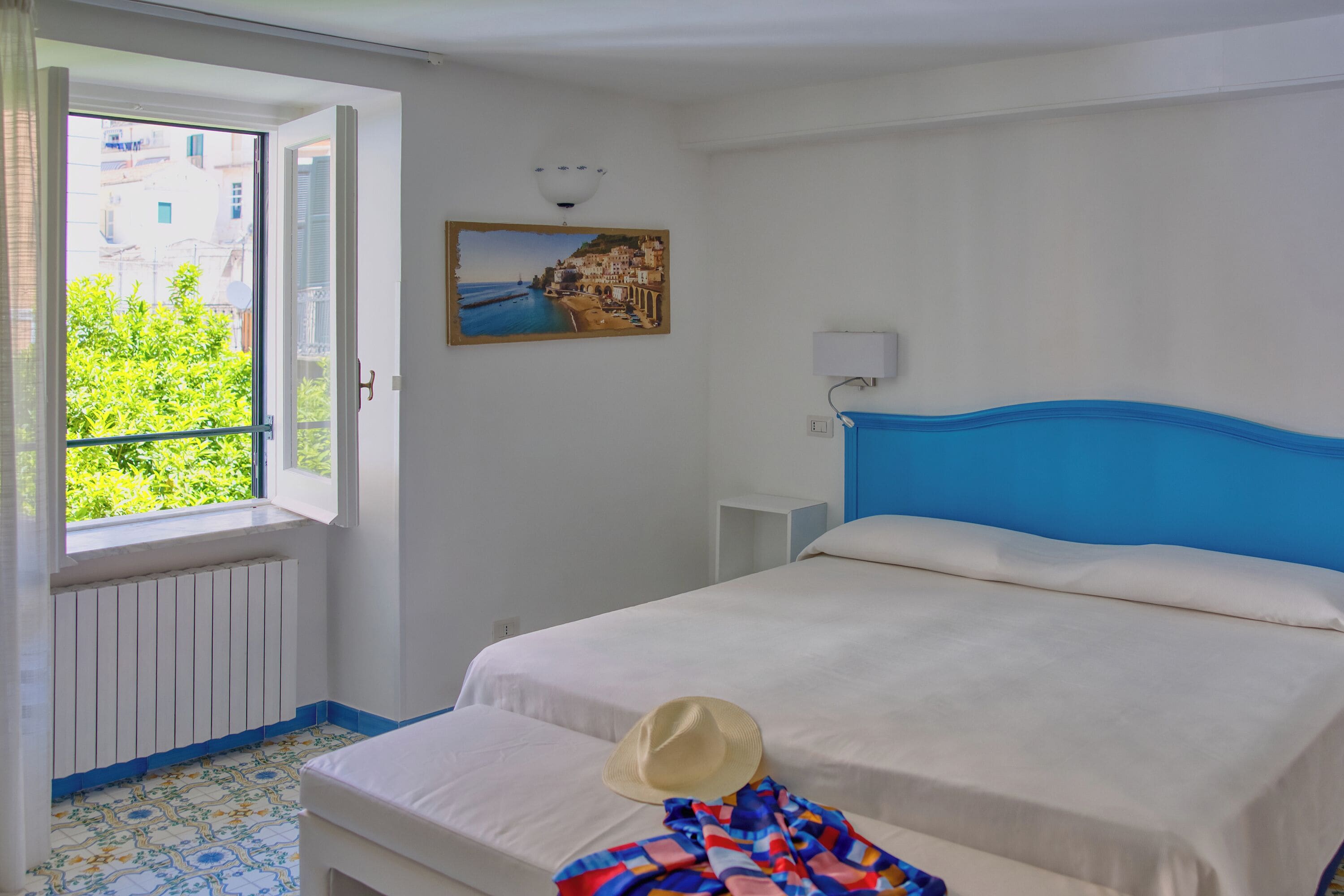A bedroom with a bed, a window with a view of greenery, a painting on the wall, and a radiator.