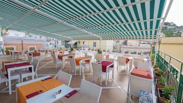 Un área de comedor al aire libre con mesas y sillas, un toldo a rayas y plantas en macetas.