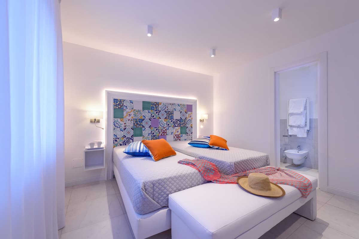 Une chambre d’hôtel moderne avec deux lits, une petite table de chevet, une salle de bain avec un lavabo et des toilettes, et une décoration murale colorée.