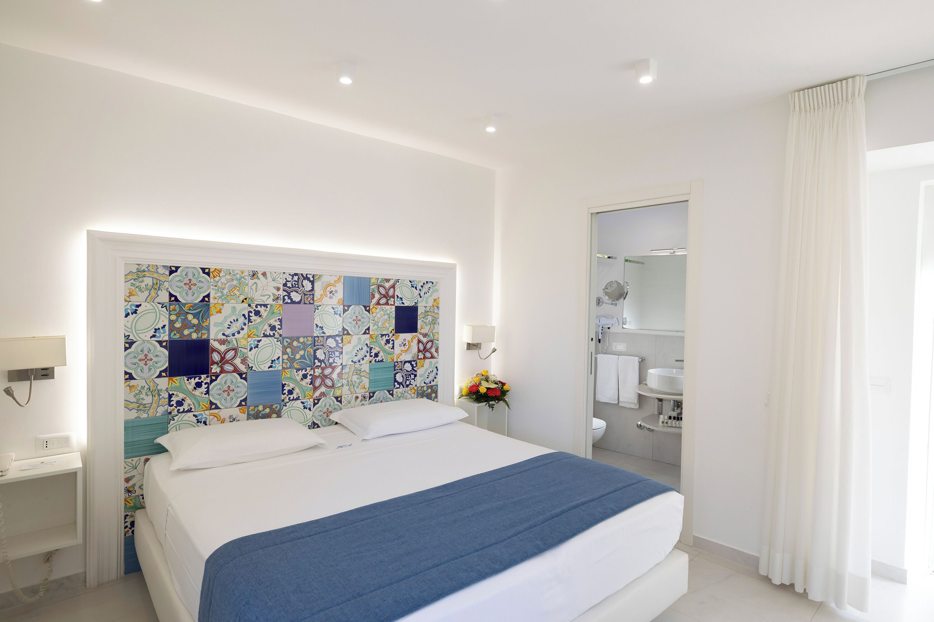 A hotel room with a bed, a bedside table, a nightstand, a bathroom with a toilet, a sink, and a mirror.