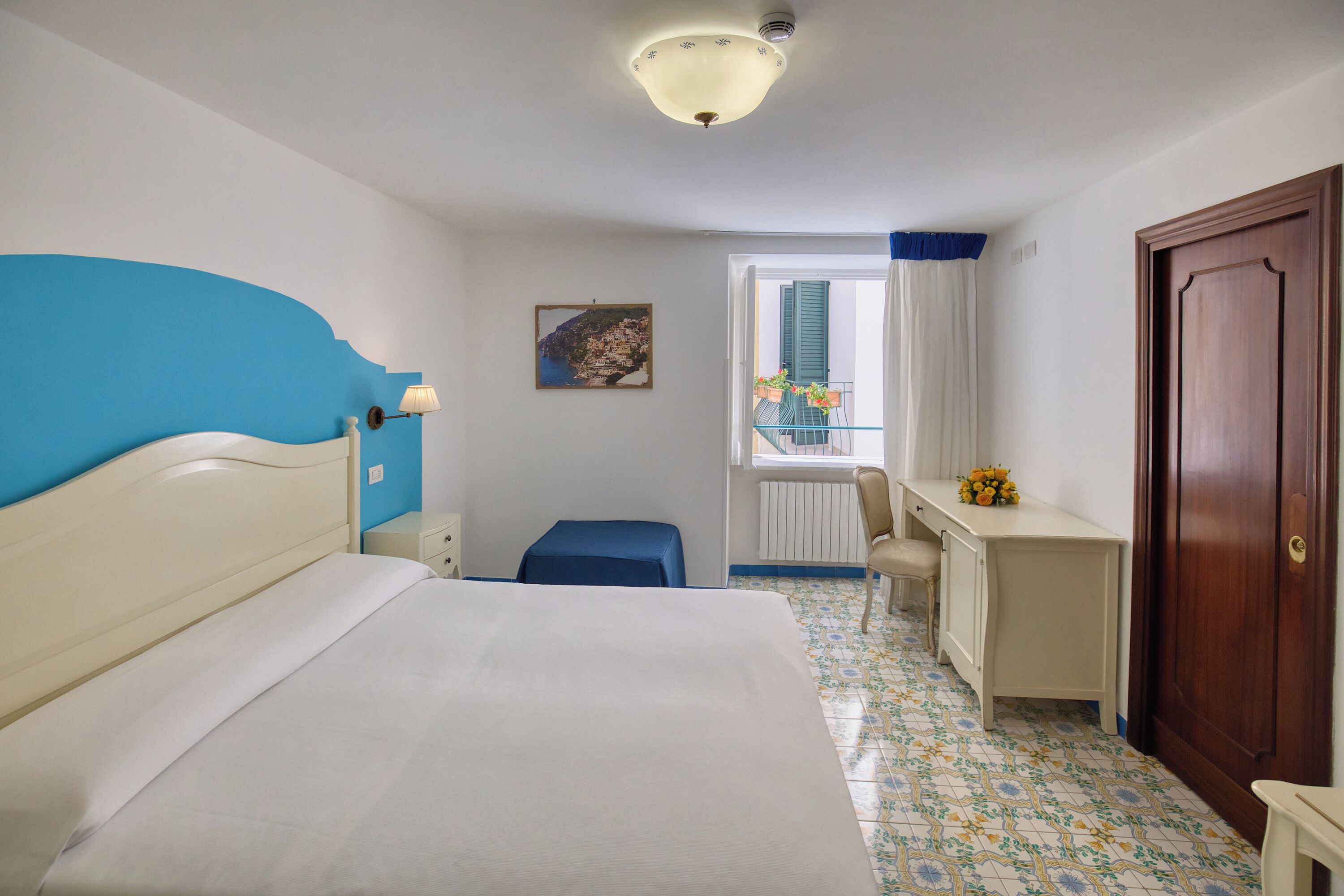 A hotel room with a bed, a desk, a chair, a window with curtains, a door, and a picture on the wall.