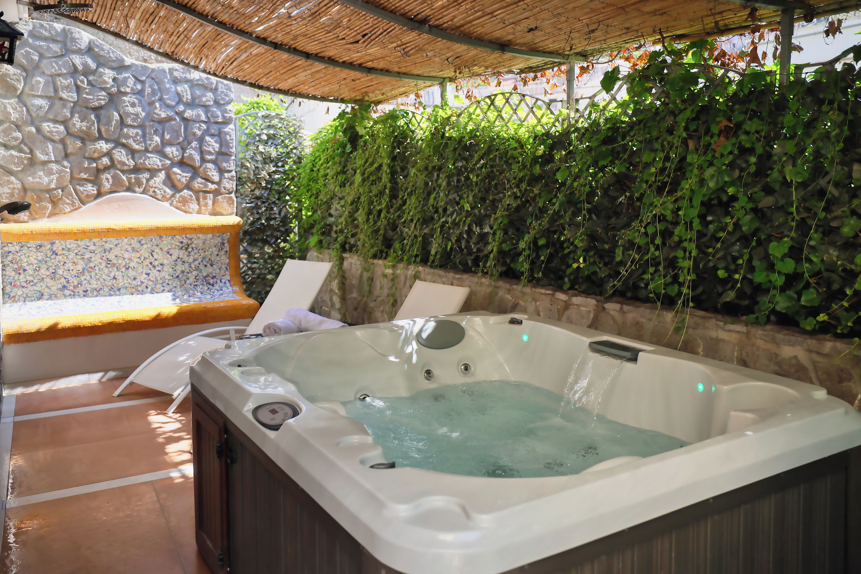 A hot tub under a pergola with a stone wall and greenery in the background.