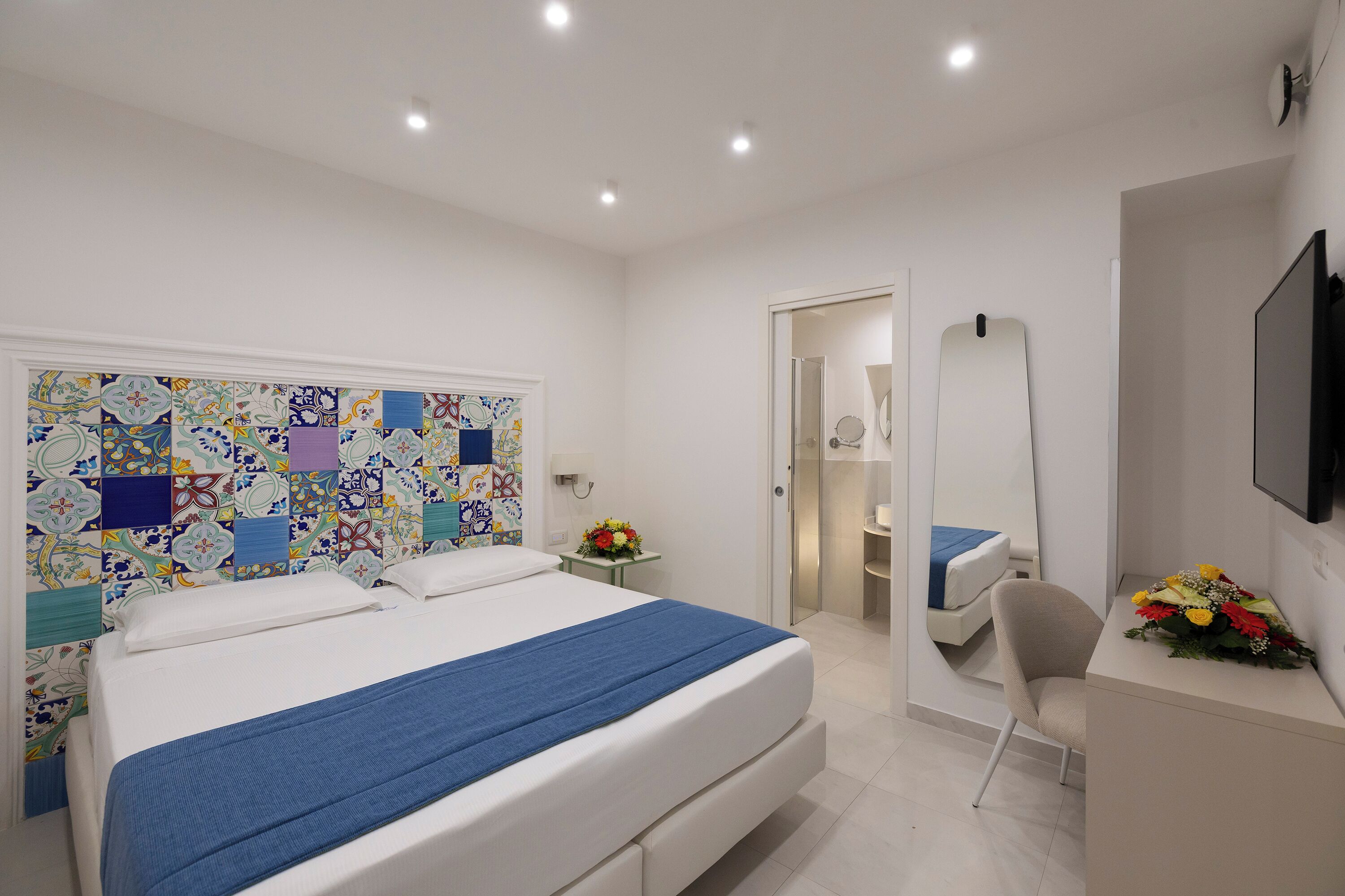 Une chambre moderne avec un grand lit, une table de chevet avec un miroir, un bureau agrémenté d’un vase de fleurs et un téléviseur fixé au mur.