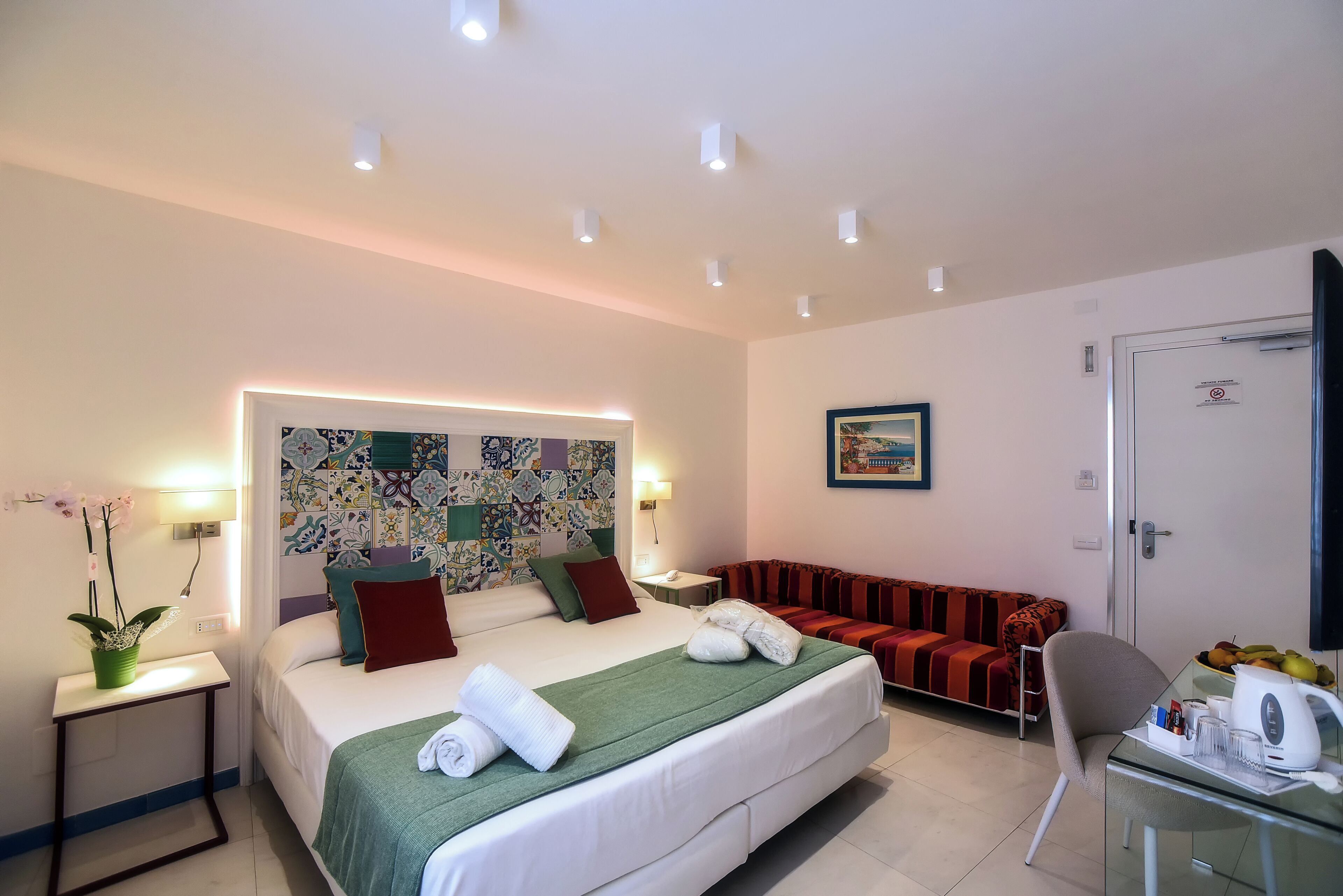 Una habitación de hotel con cama, sofá, sillón, una mesita con hervidor y una pared con estampado colorido.