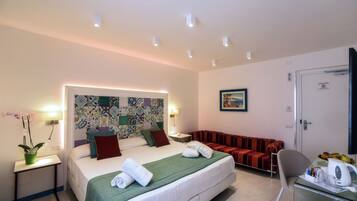 Una habitación de hotel con cama, sofá, sillón, una mesita con hervidor y una pared con estampado colorido.