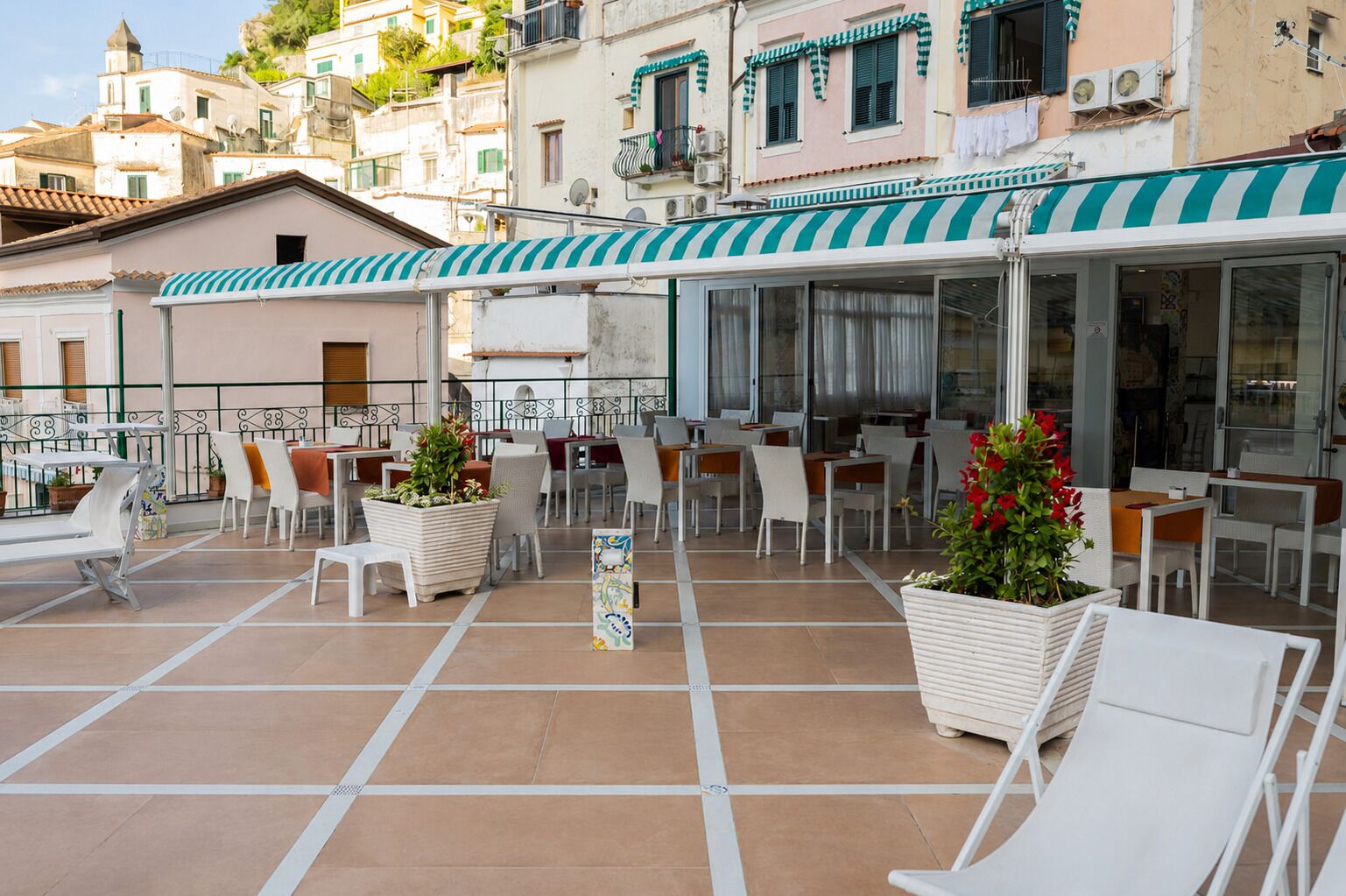 Un café al aire libre con sillas y mesas blancas, plantas en macetas y un toldo a rayas.