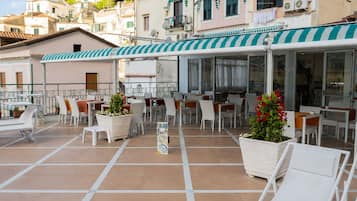 Un café al aire libre con sillas y mesas blancas, plantas en macetas y un toldo a rayas.
