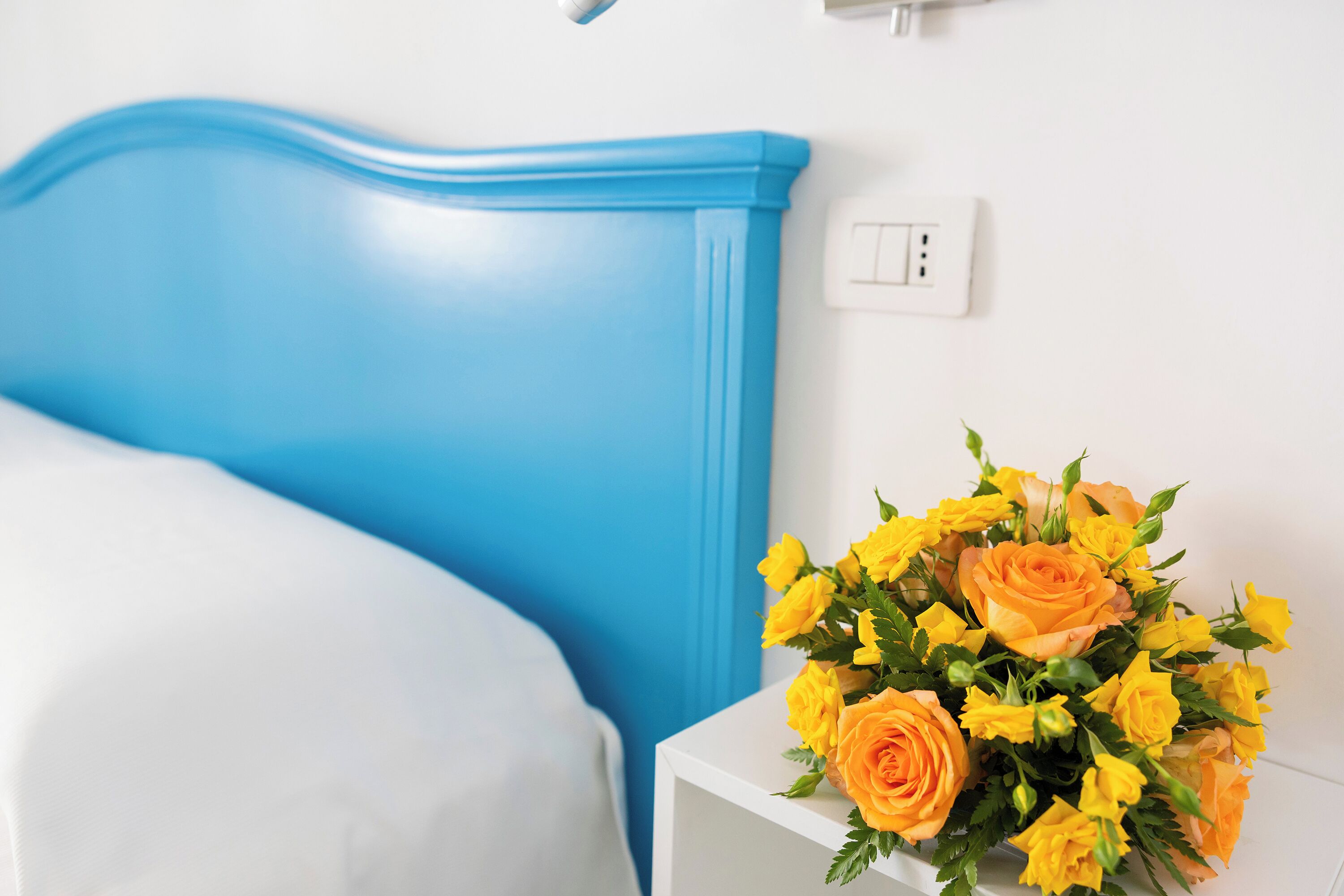 Ein Bett mit blauem Kopfteil, ein Nachttisch mit einem Strauß gelber Blumen und eine weiße Wand mit einem Lichtschalter.