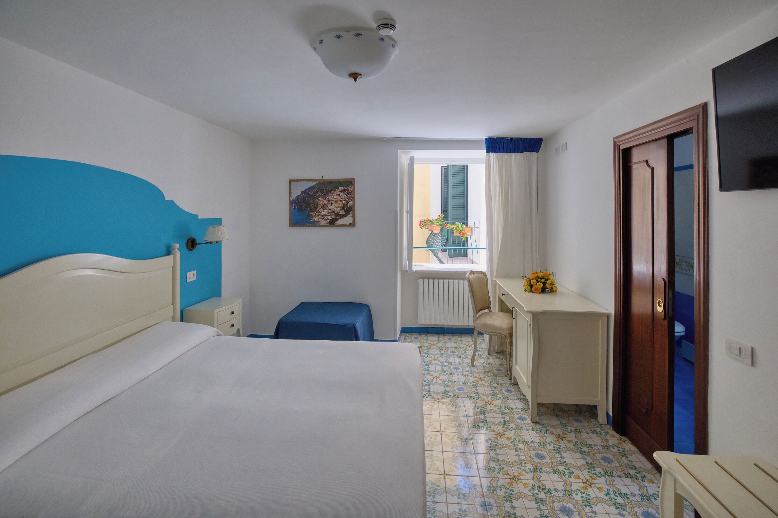 A bedroom with a bed, a desk, a chair, a window with a view, and a door.