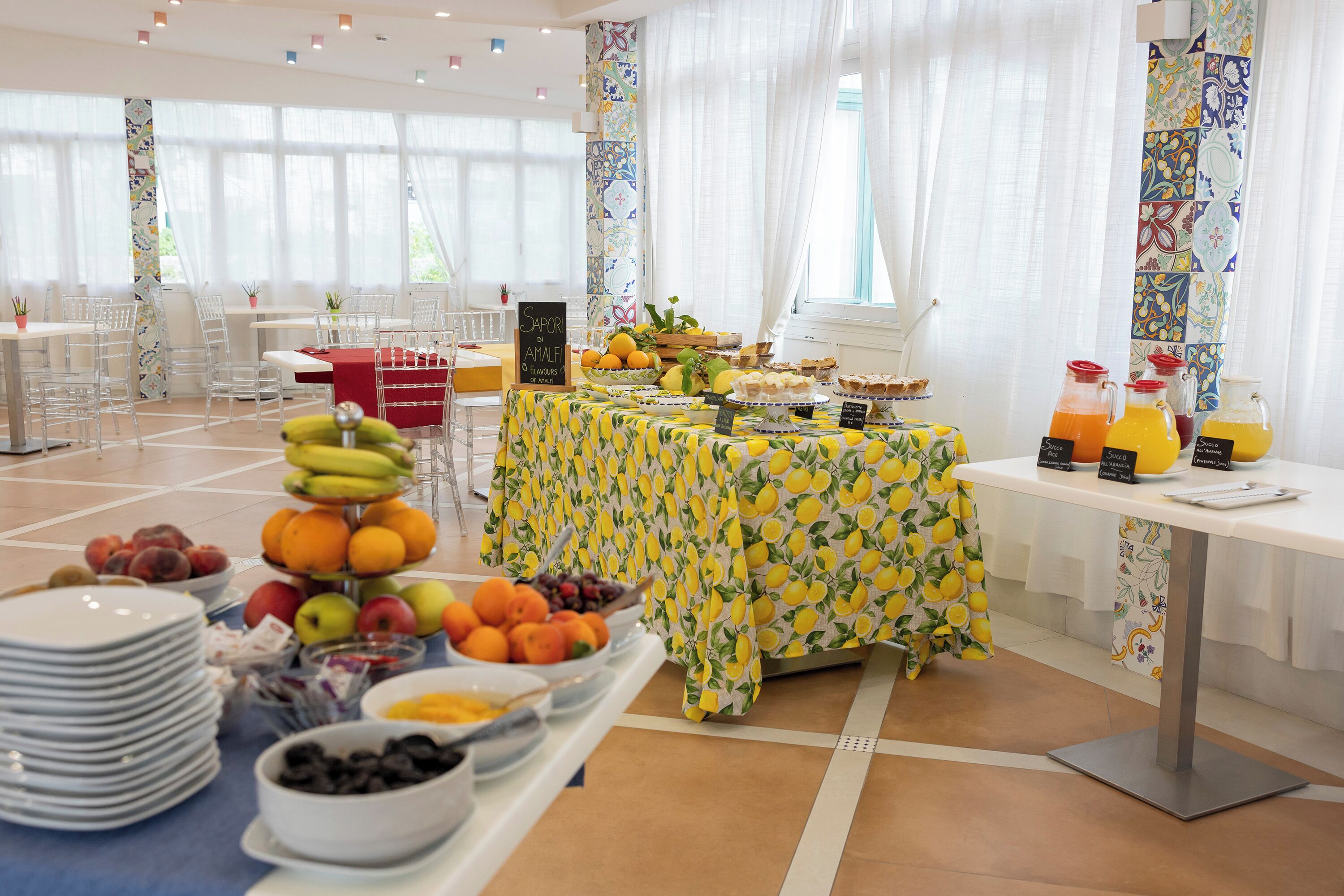 A buffet setup with various fruits and beverages, a table with a lemon pattern tablecloth, and a view of a bright room with large windows and curtains.