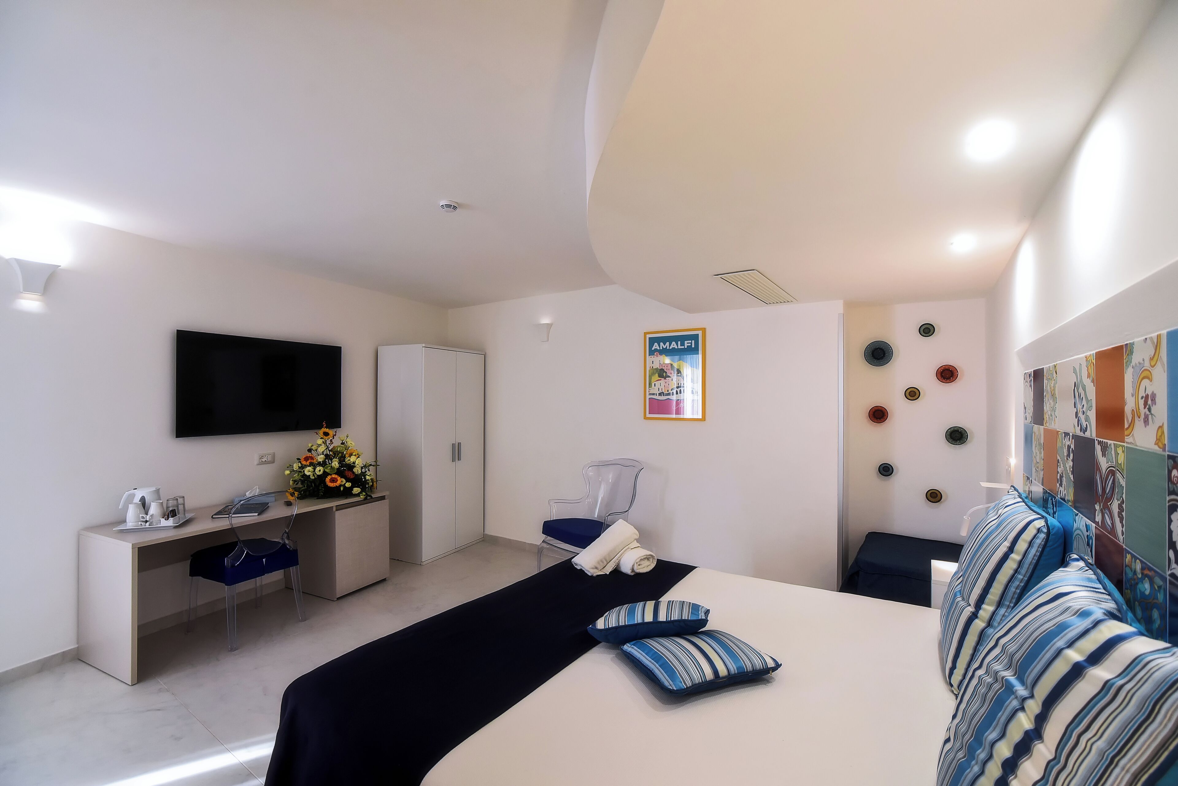 A modern hotel room with a bed, a desk, a chair, a TV, and a poster on the wall.