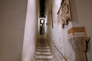 Un escalier étroit doté d’une main courante, menant à une pièce éclairée par un luminaire et où est affiché un panneau indiquant une direction.