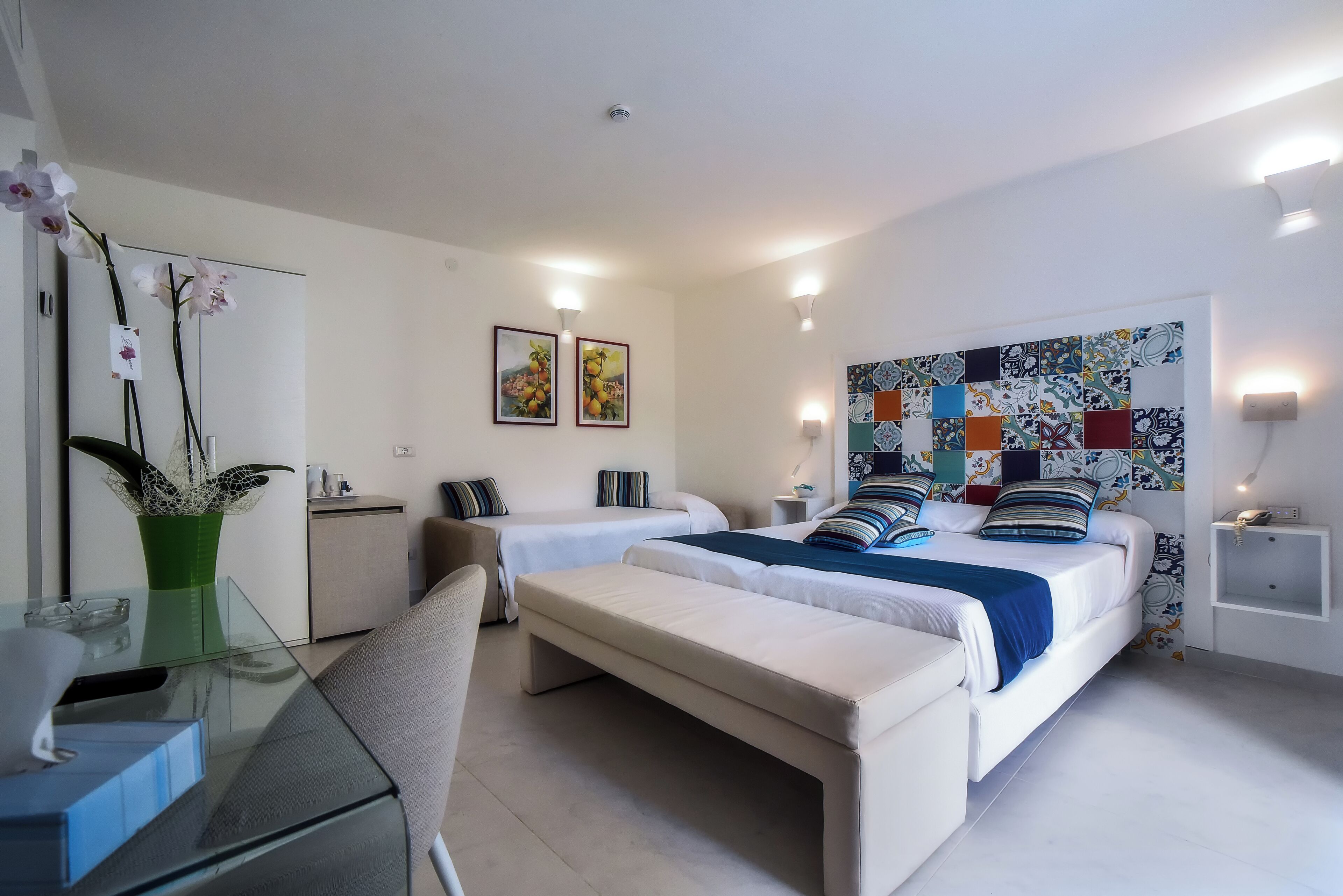 A hotel room with two beds, a desk, a chair, a bench, a small cabinet, a vase with flowers, and wall art.