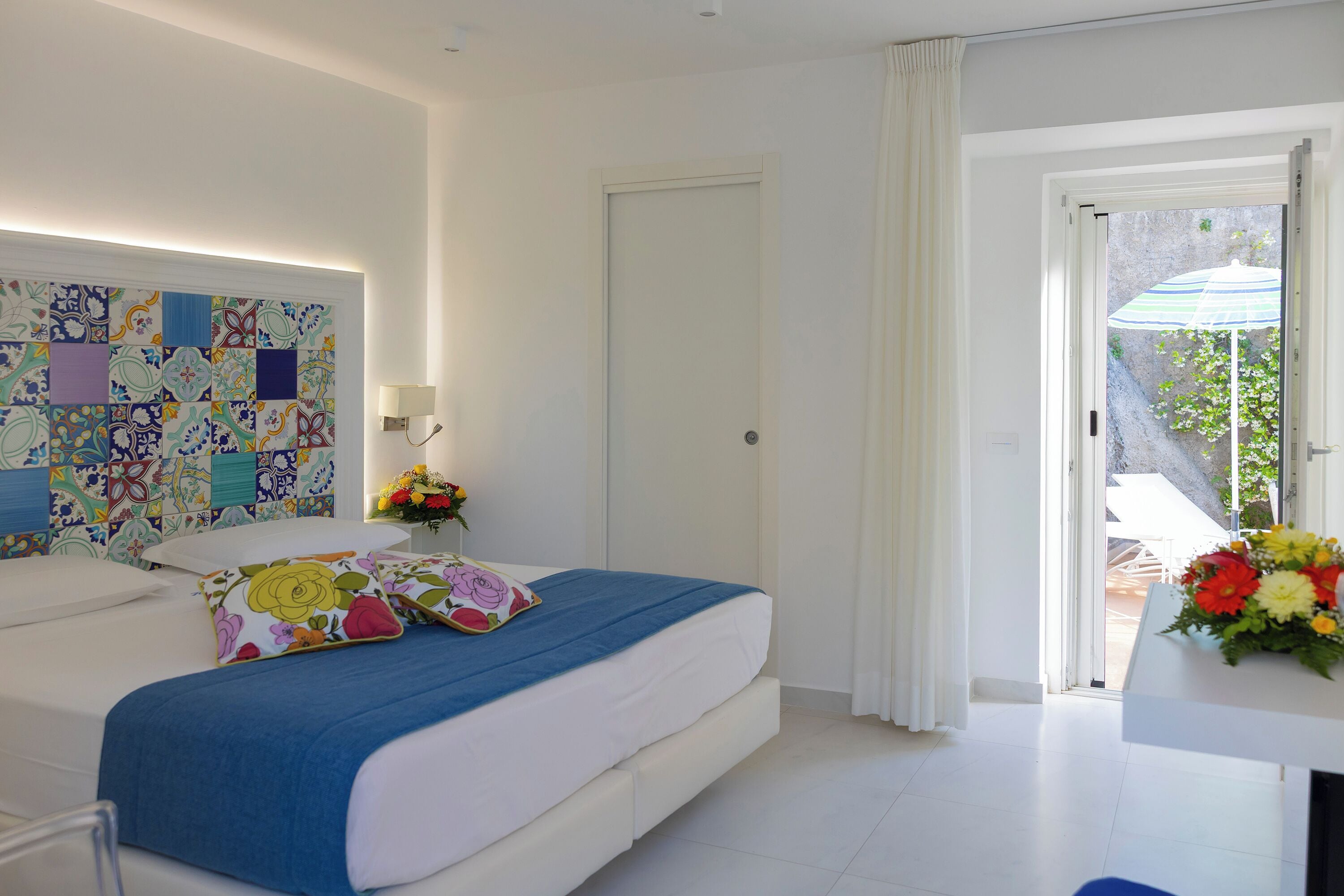 A bedroom with a bed, a nightstand, a wall with a colorful pattern, a door, and a view of the outdoors.
