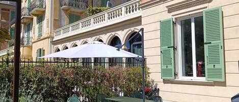 A courtyard with outdoor seating, a white umbrella, and a building with green shutters.