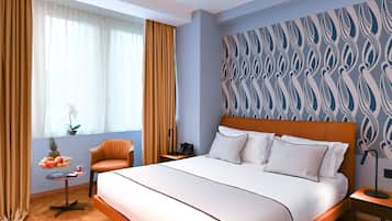 A hotel room with a bed, a chair, a small table with fruit, a nightstand, and a patterned wall.