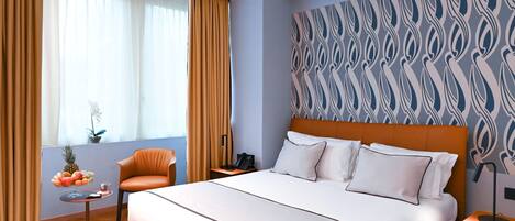 Habitación de hotel con cama, una silla, una mesita con fruta, una mesita de noche y pared con diseño.