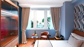 A hotel room with a bed, a chair, a small table with fruit, a phone on a side table, and a window with curtains.