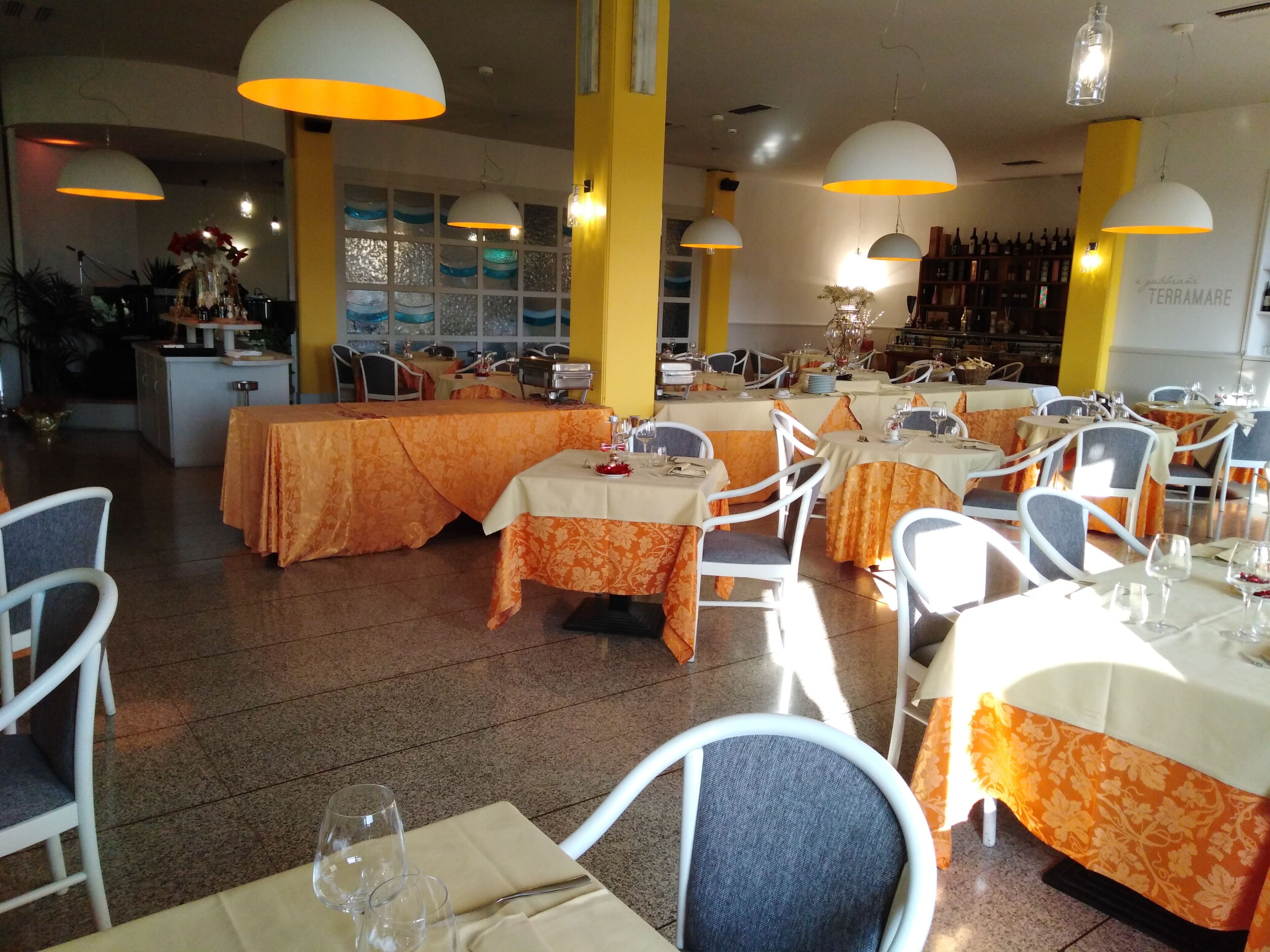 Un restaurante con varias mesas y sillas, cada una cubierta con un mantel. El espacio tiene piso de baldosas e iluminación con lámparas colgantes.