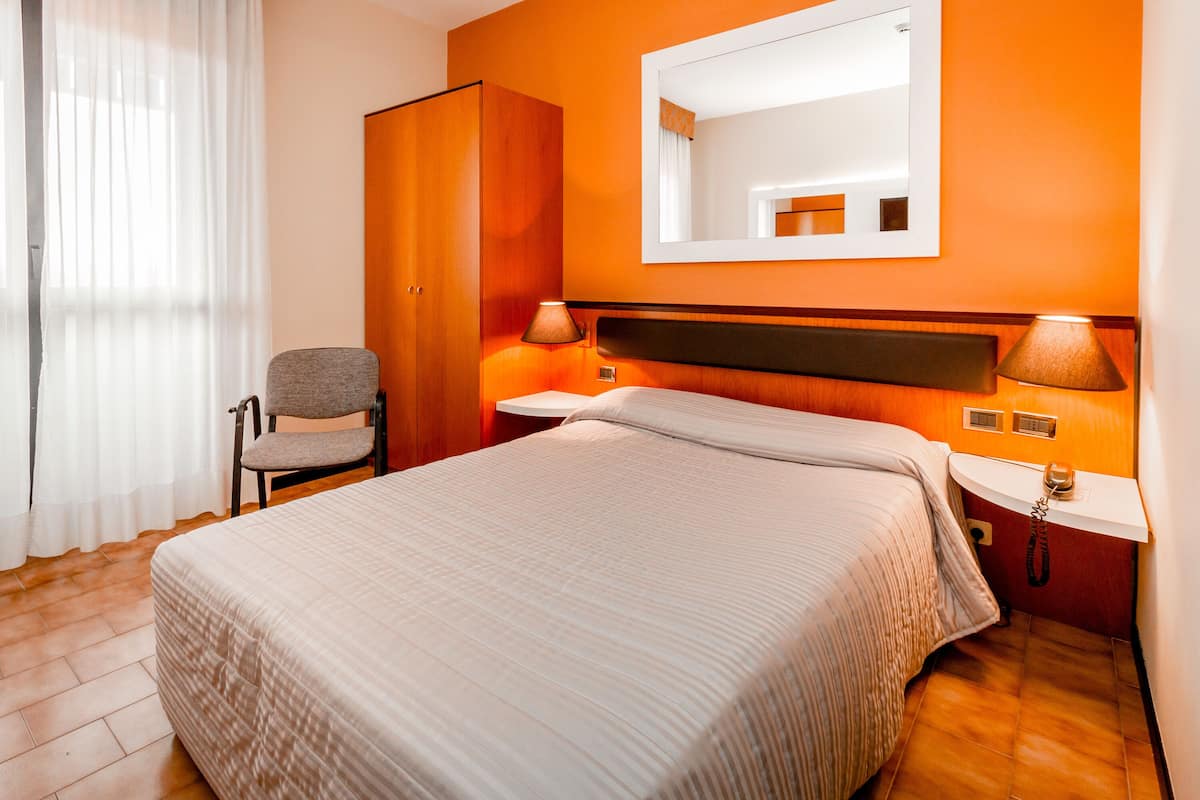Une chambre d’hôtel avec un lit, une chaise, une armoire, une table de chevet avec une lampe et un miroir au mur.