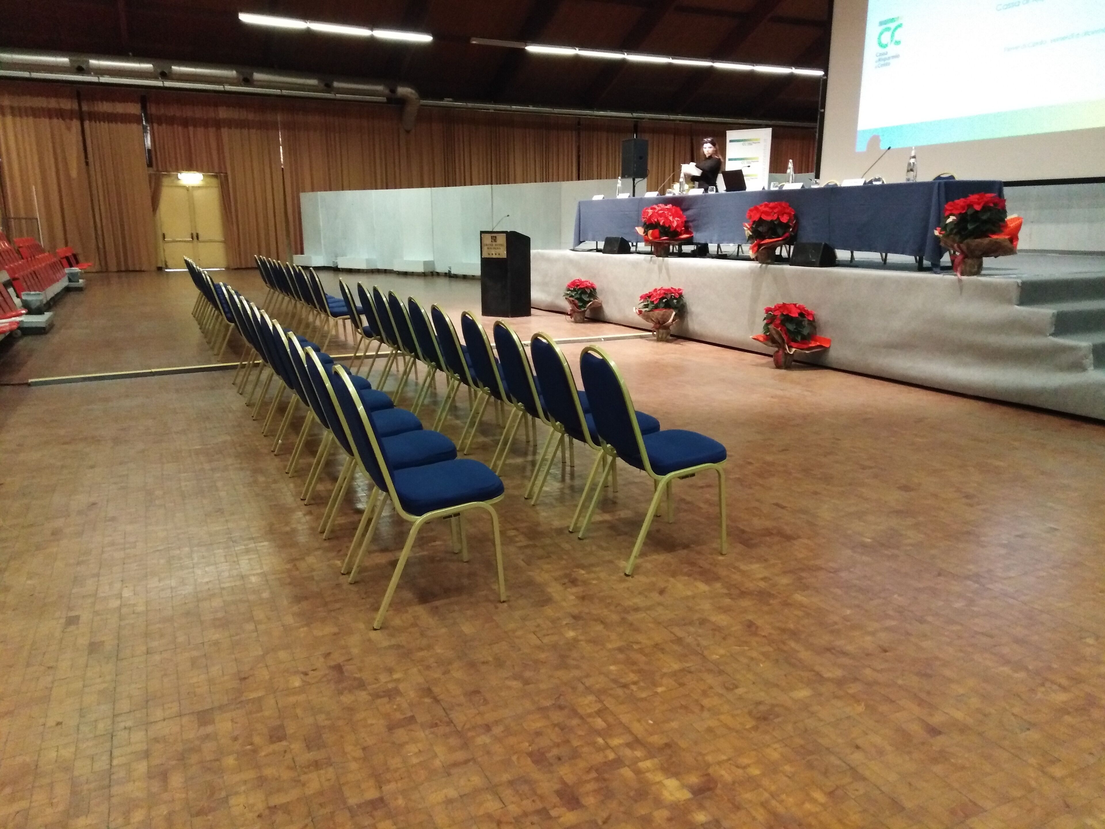 Un salón de conferencias con filas de sillas azules, un escenario con un atril y arreglos florales rojos.