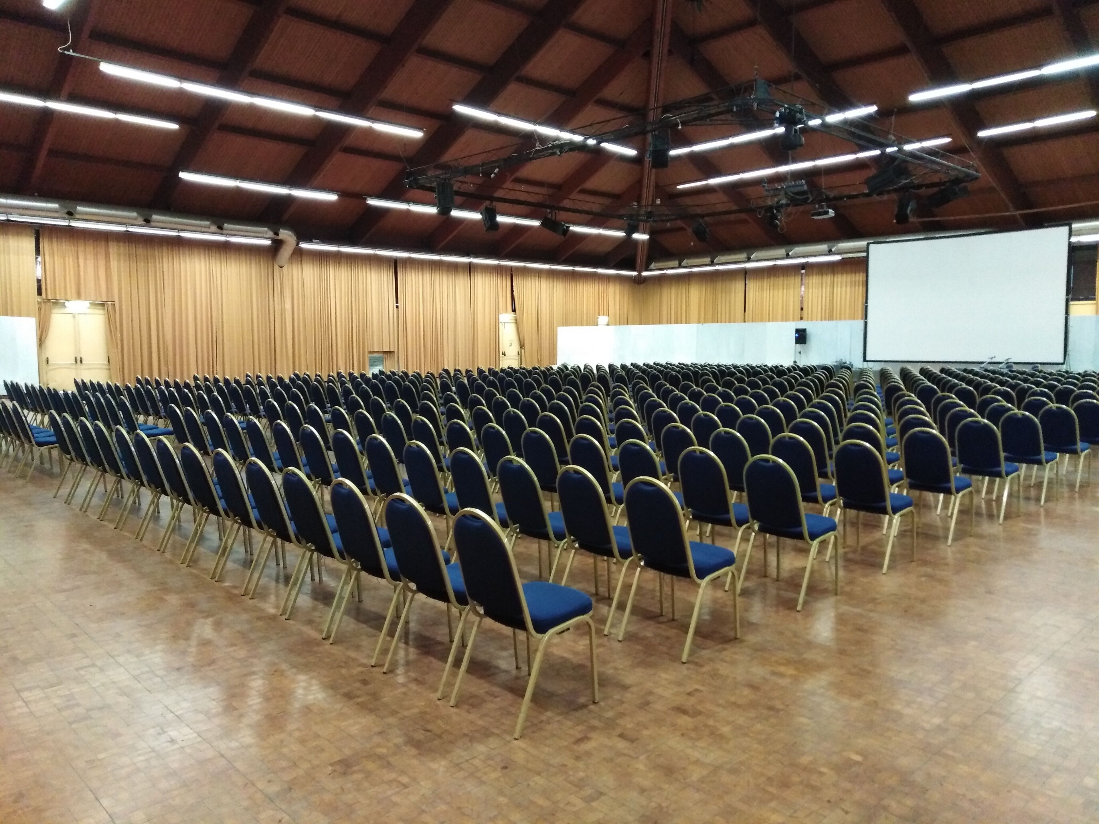 Een ruime zaal met rijen blauwe stoelen en een wit scherm aan de voorzijde.