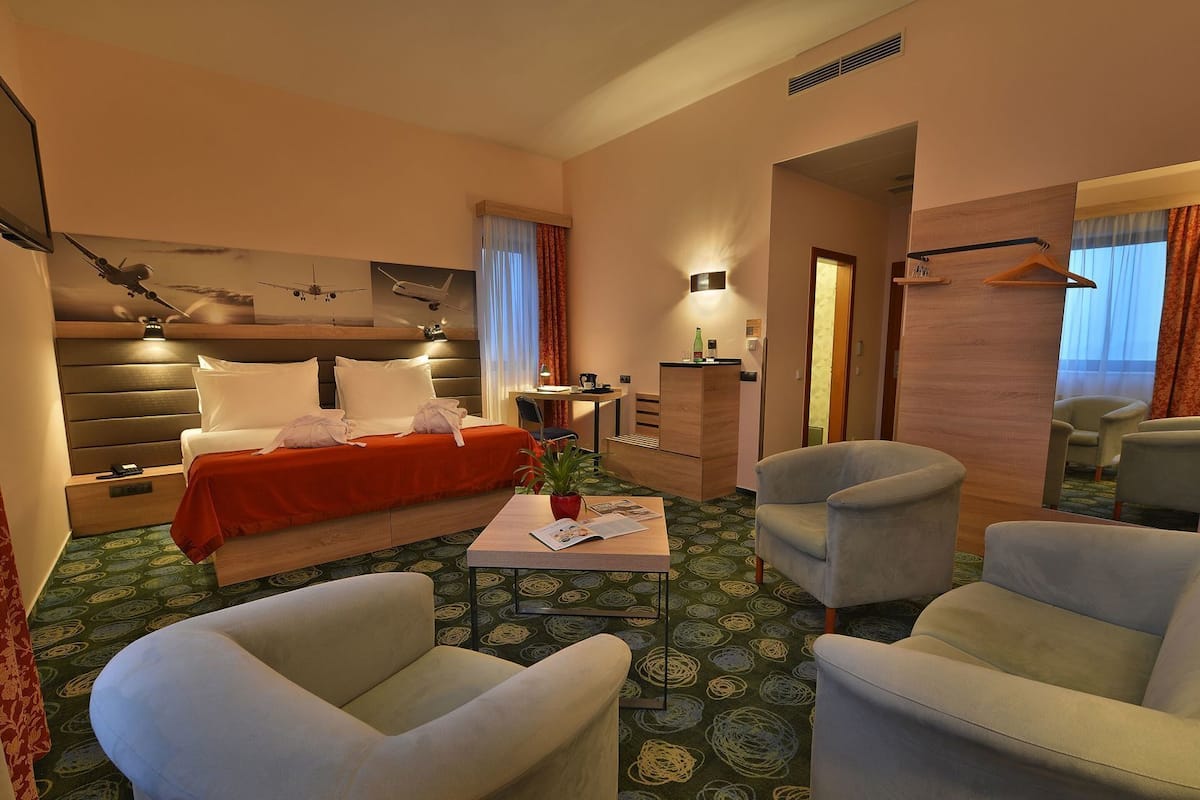 A hotel room with a bed, two armchairs, a small table, a desk, a mirror, and a wall-mounted artwork.