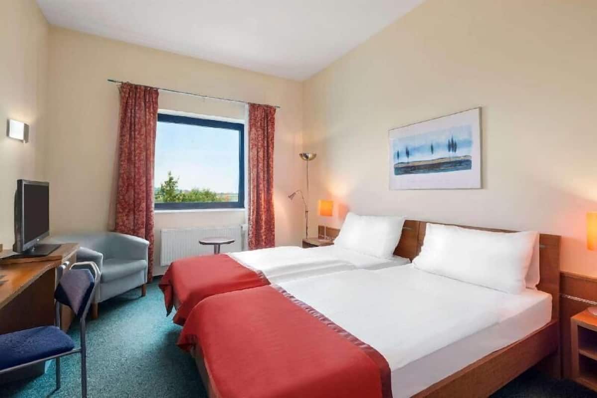 A hotel room with two beds, a desk with a chair, a TV, a window with curtains, a floor lamp, and a painting on the wall.