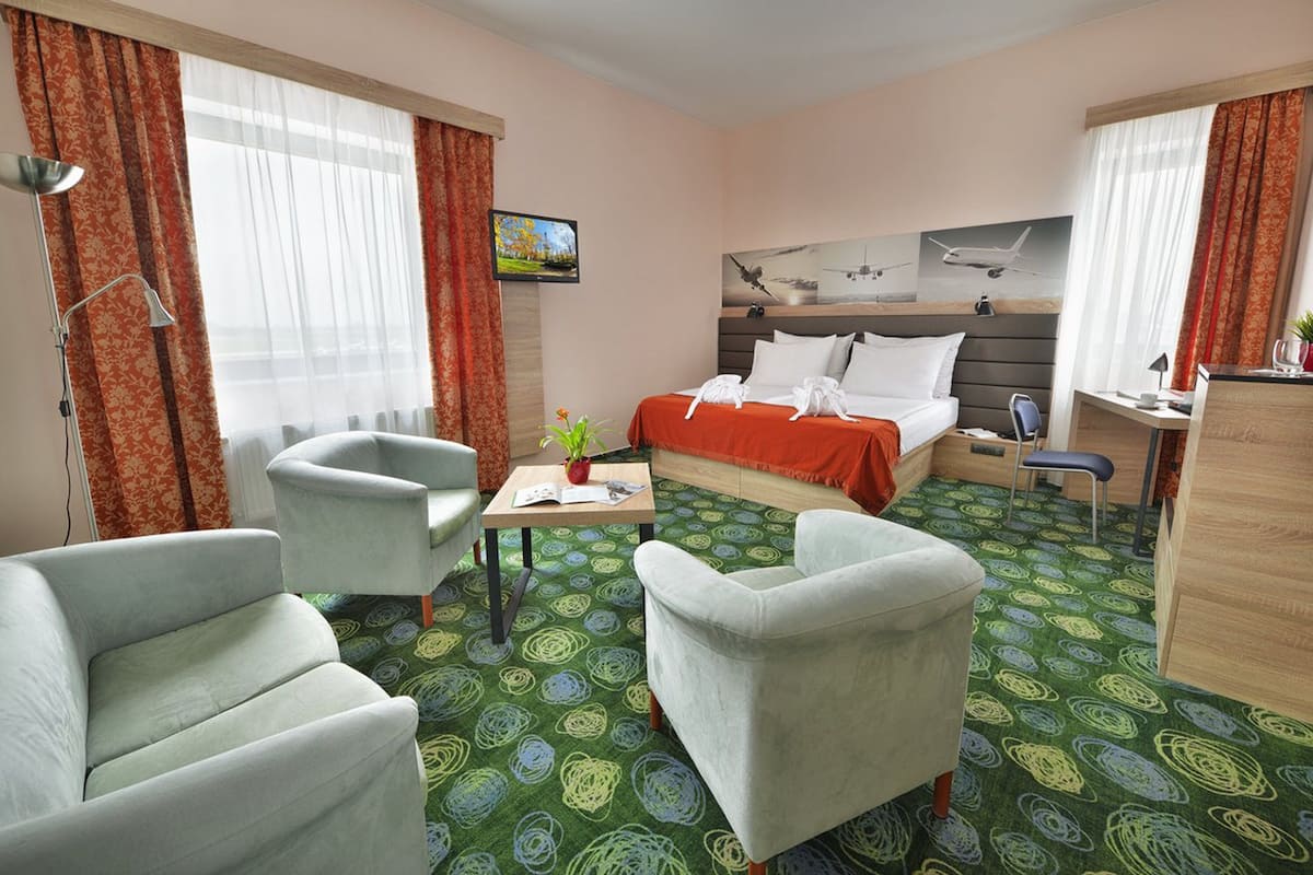 A hotel room with a bed, two armchairs, a desk, a TV, and a window with curtains.