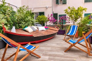 Un área de descanso con forma de barco de madera, con cojines, rodeada de plantas en macetas y sillas a rayas azules.