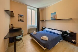 Habitación de hotel con cama, escritorio, silla, ventana y estante en la pared.