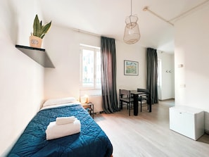 A bedroom with a bed, a desk, a chair, a small table, a plant, and a hanging light fixture. - Hotel Britannia (Genoa)