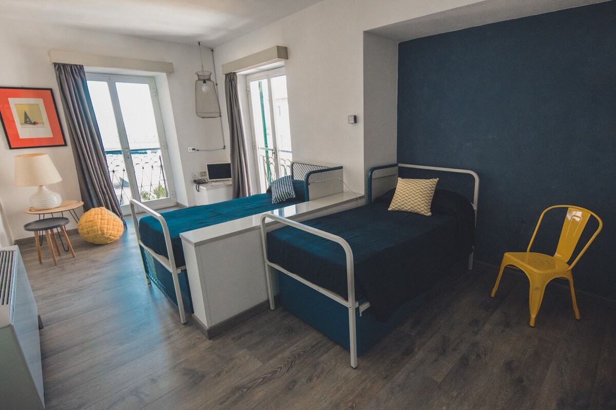A compact bedroom with a single bed, a small desk, a chair, and a view of the outdoors.