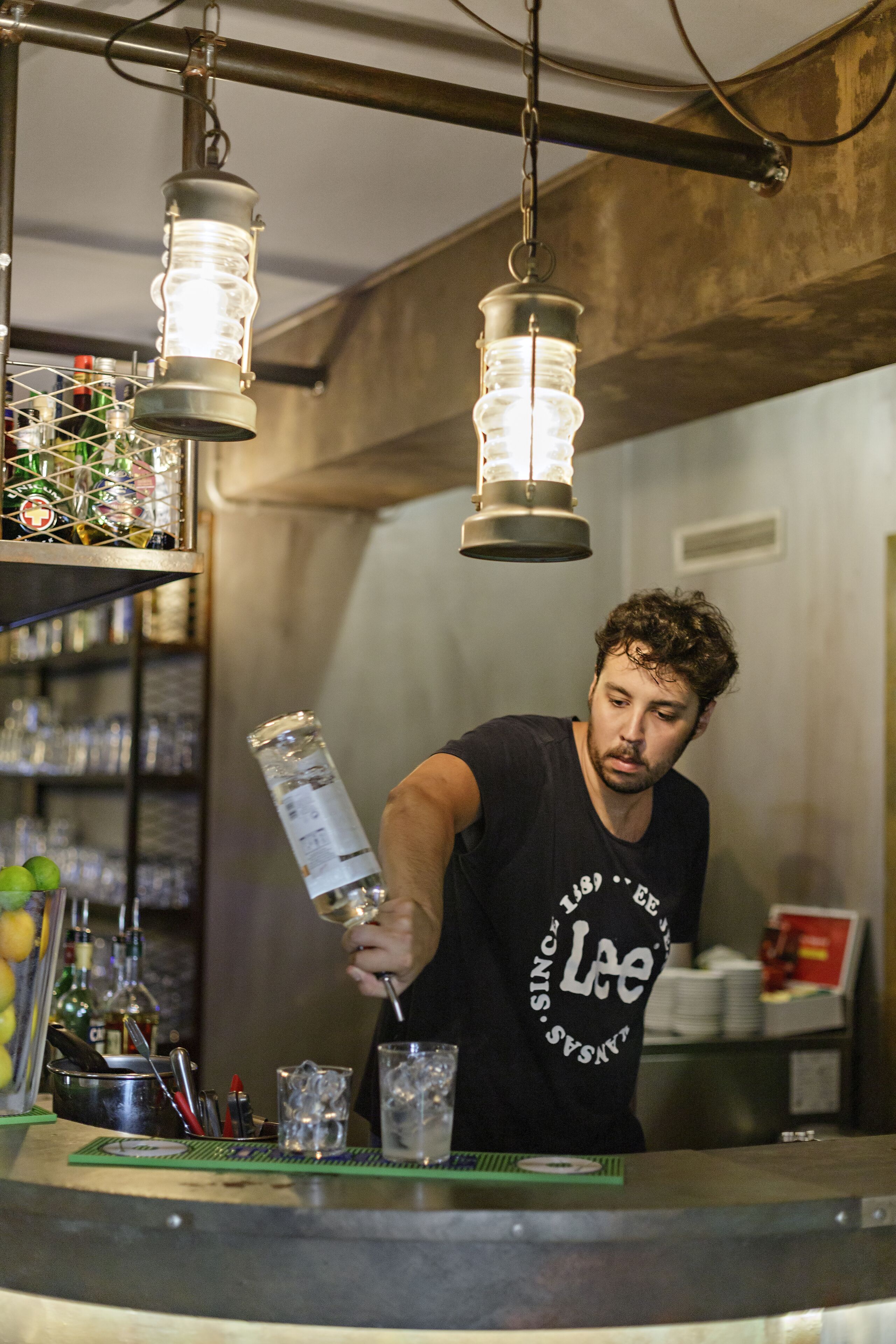 Un barman prépare un cocktail dans un bar décoré de guirlandes lumineuses et doté d’étagères remplies de bouteilles.