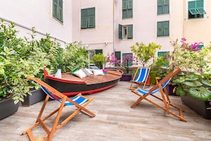An outdoor seating area with wooden chairs and a boat-shaped table, surrounded by potted plants and a building with green shutters.