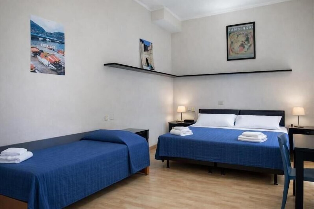 A hotel room with two single beds, a desk, and a chair. There are wall-mounted shelves with artwork and a framed picture above the beds.