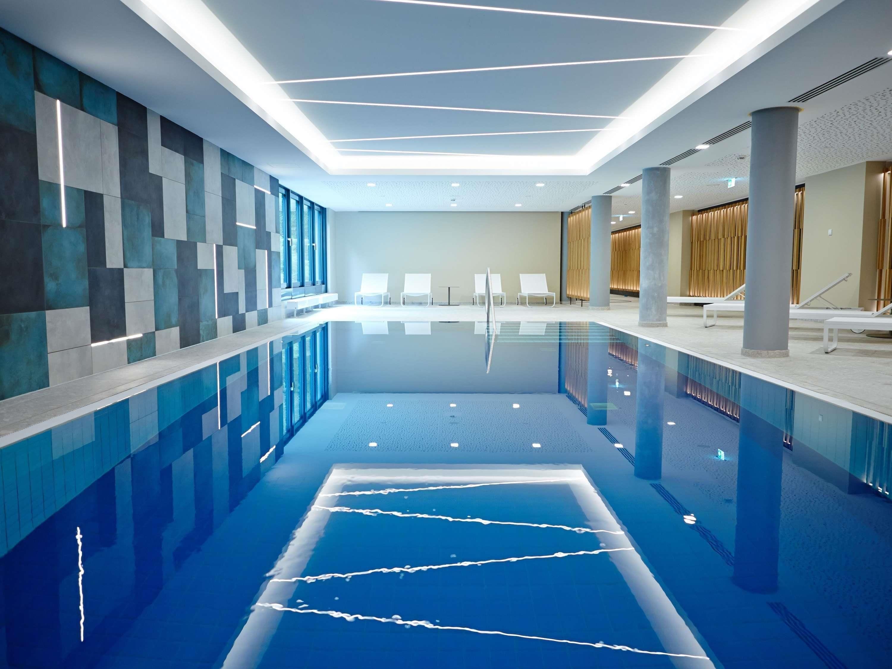 Una piscina interior moderna con agua cristalina, sillas de descanso blancas y un diseño de pared geométrico.