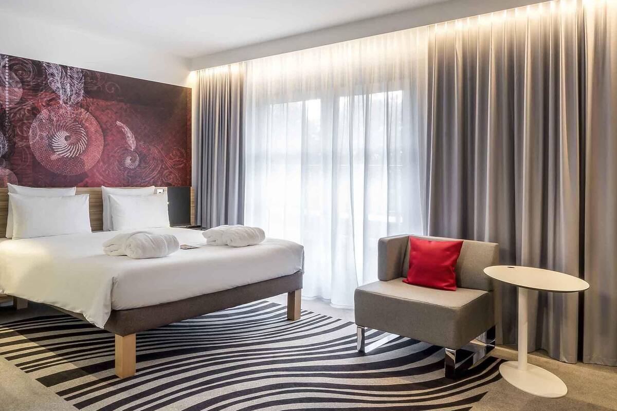 Ein modernes Hotelzimmer mit einem großen Bett, einer kleinen Sitzecke, einem runden Tisch und einem großen Fenster mit Vorhängen.