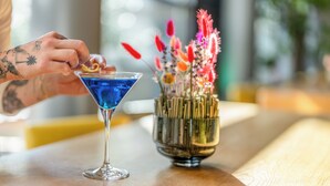 A martini glass with a blue liquid on a wooden table next to a decorative vase with various flowers. - Novotel Muenchen City (Munich)
